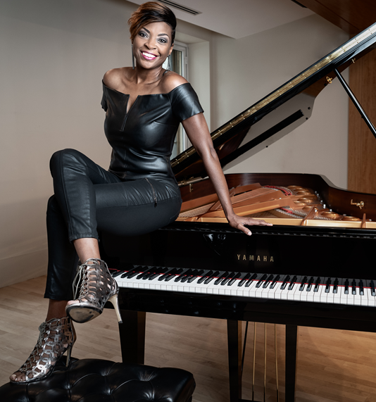 Renowned Black artist, Jade Simmons, sits on a black grand piano, smiling at the camera. She's wearing a black fitted jumpsuit with black lace-up heels.