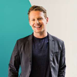 Eric Termunde. Headshot of Eric Termunde smiling, wearing a black jacket and black T-shirt, posing in front of a turquoise and white wall.