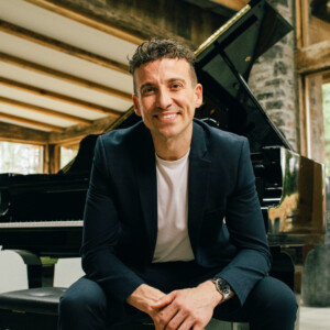 Cameron Atlas. Headshot of Cameron Atlas wearing a black suit over a white t-shirt. He is leaning forward on his elbows and sitting at a piano.