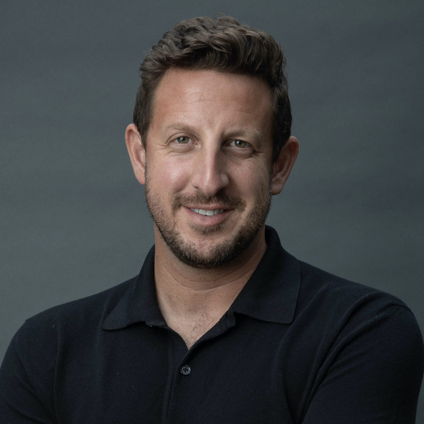 Zack Kass Headshot. A portrait shot of Zack Kass smiling in front of a dark grey background and wearing a black polo shirt.