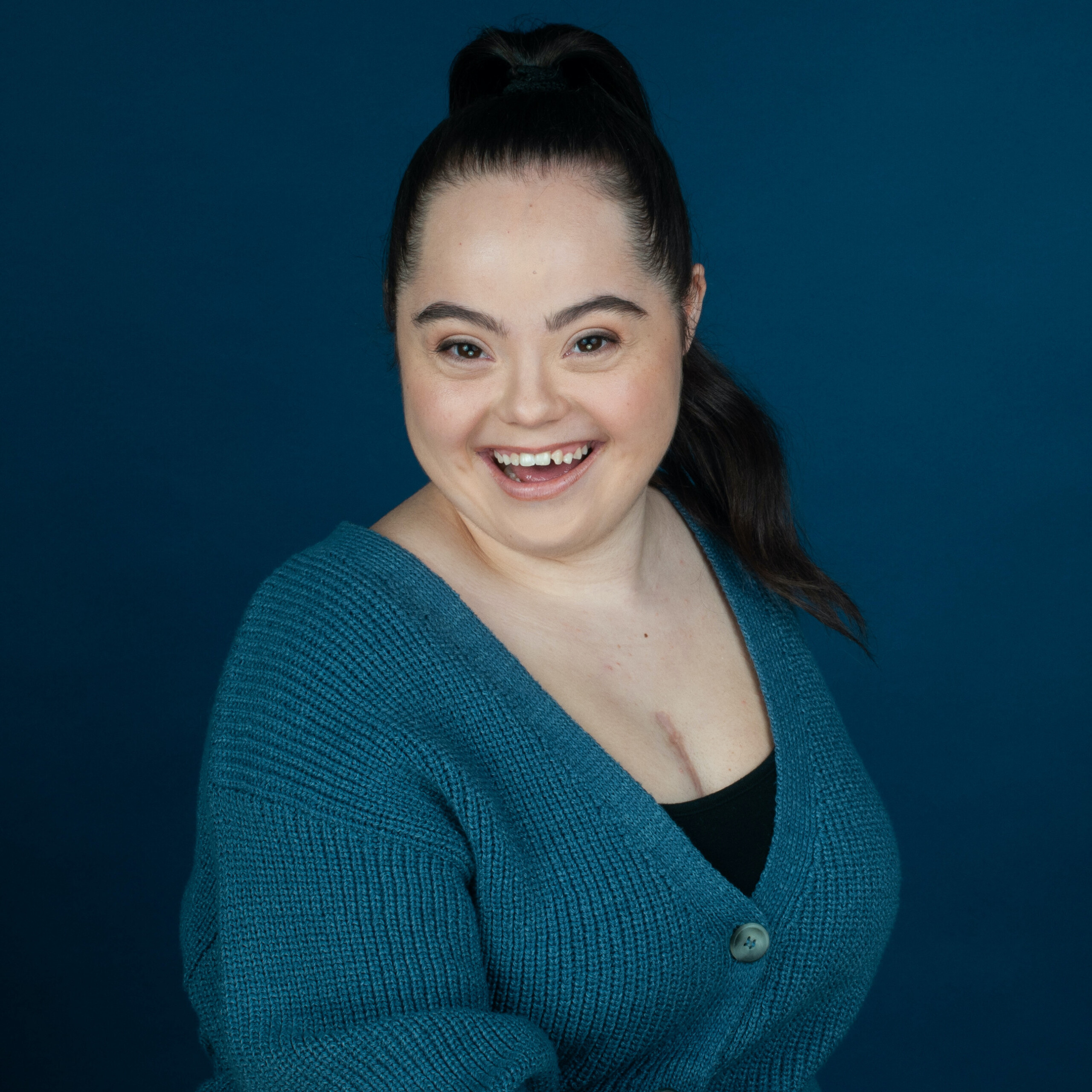 Headshot of Madison Tevlin smiling in front of a dark blue backdrop. Her hair is up in a ponytail and she's wearing blue cardigan over a black top.