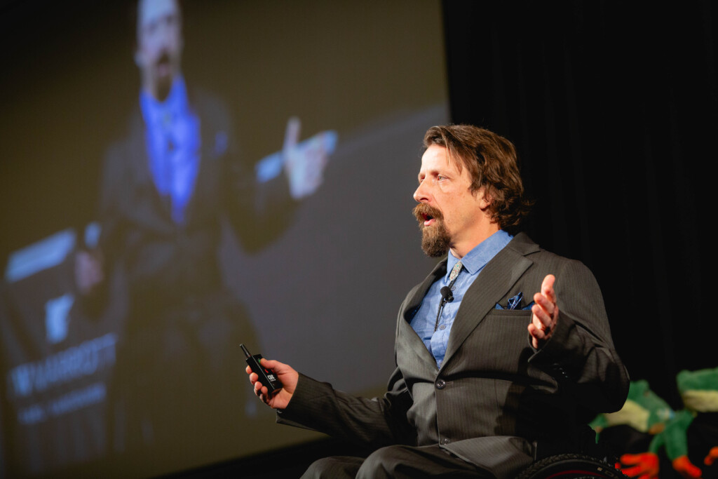 Josh Dueck, wearing a black suit and blue undershirt, sitting on stage delivering a keynote. His hands are open in front of him and in the background in a large screen projecting a live feed of him presenting.