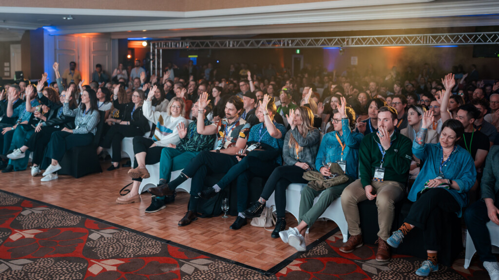 A large conference room filled with an eager and excited audience and everyone raising a hand in the air. The room glows in war stage lights illuminating the audiences' faces.