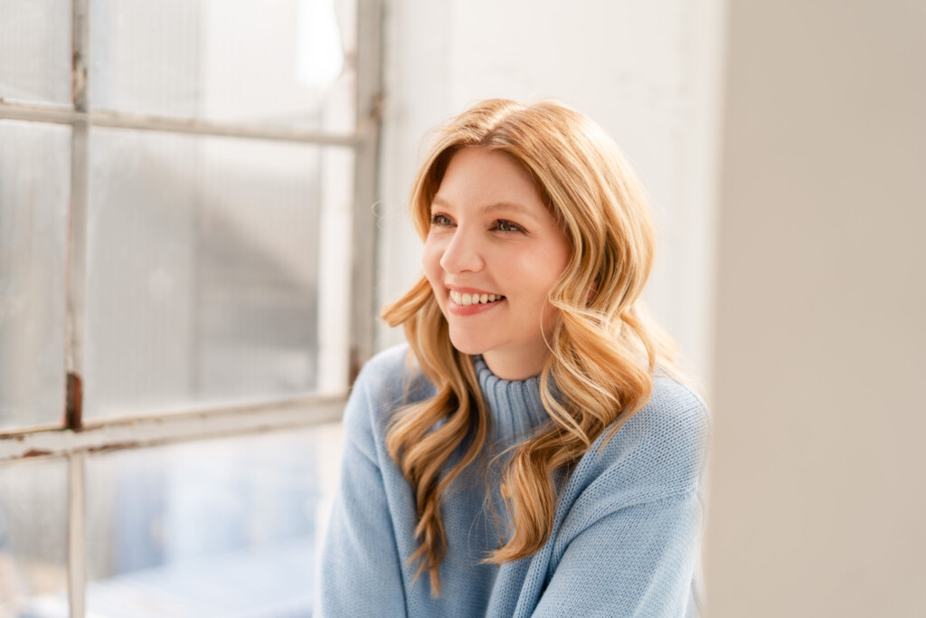 Jessica Moorhouse headshot of her sitting by a light-filled window, smiling and gazing away from the camera. She's wearing a light blue blouse with her blonde hair down and curled.