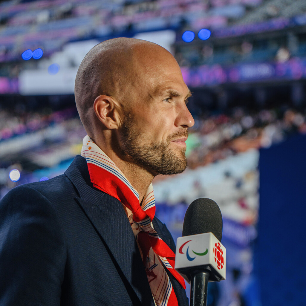 Devin Heroux. Photo of Devin Heroux with a microphone wearing a dark navy jacket.