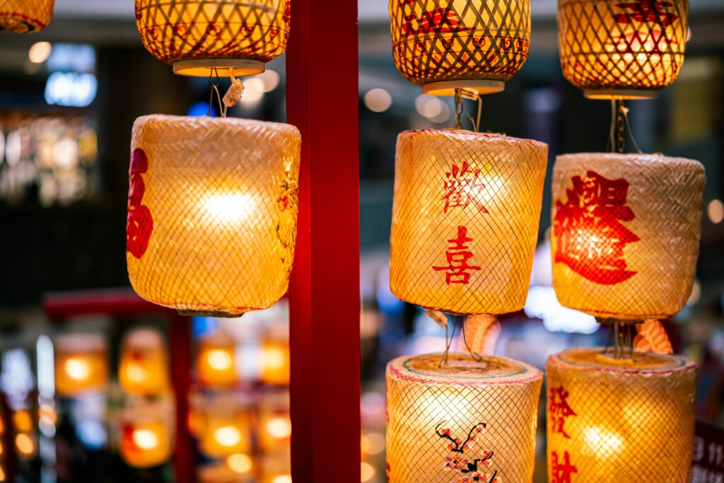 Lit Asian lanterns hanging vertically and flowing with a warm light. There are Chinese characters printed on the outside of the lanterns in red font.