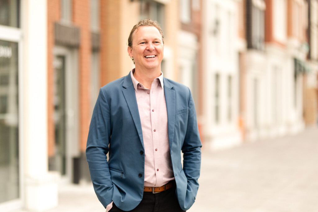 Headshot of Doug Griffiths keynote speaker. Doug is standing outside, smiling, with his hands in his pant pockets. He is wearing a light pink button up underneath a dark blue blazer.