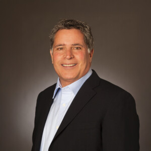 Bill Taylor. Headshot of Bill Taylor wearing a black suit jacket over a light blue collared shirt. He's posing in front of a dark brown studio background.