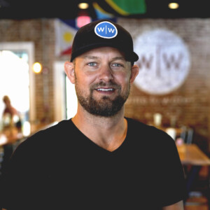 Doc Hendley. Headshot of Doc Hendley wearing a black t-shirt and a black cap with a blue and white w/w logo on it. He's posing in front of a blurred background.