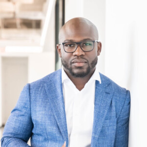 Justin Forsett. Headshot of Justin Forsett wearing a blue jacket over a white collared shirt and black rimmed glasses. He is posing in front of a bright blurred background.