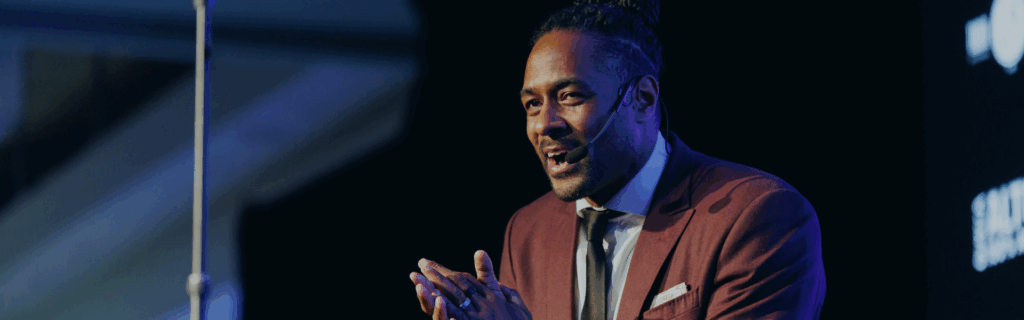 Close up photo of Jesse Lipscombe on-stage hosting the 50 Best Bars event. He is wearing a microphone and is wearing a maroon suit.
