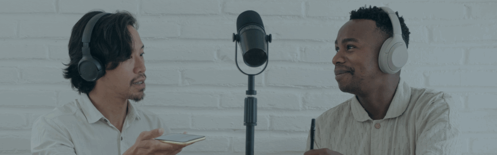 Two males sitting across from each other conversing. Both are wearing headphones and a podcast microphone is between them.