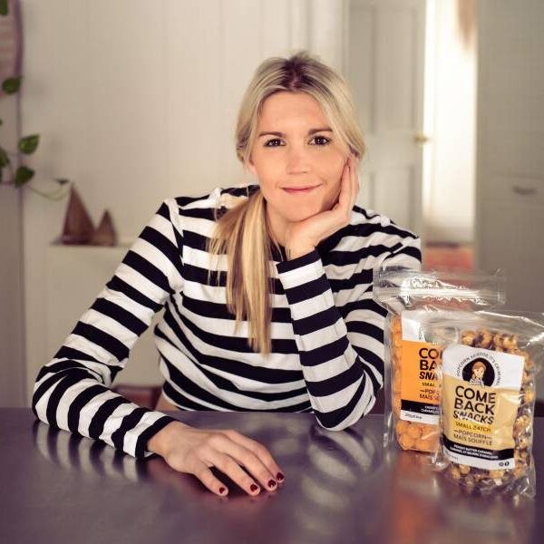 Photo of keynote speaker, Emily O'Brien, smiling, and sitting at a kitchen table. She is wearing a striped, black and white long-sleeve shirt. Two bags of her Comeback Snacks are on the table in front of her.