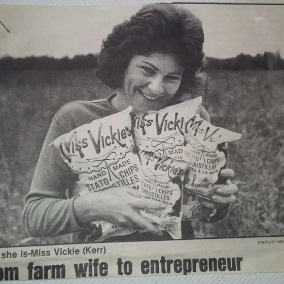 Black and white newspaper clipping of founder and creator of Miss Vickie's, Vickie Kerr. Vickie is holding 4 bags of Miss Vickie's chips in her arms.