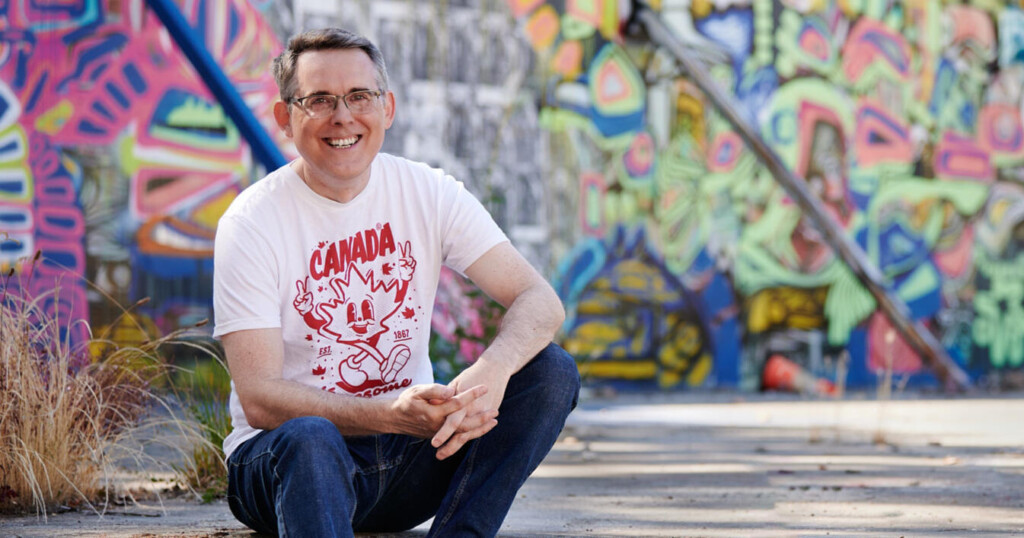 Image of Tod Maffin wearing a white and red Canada graphic t-shirt sitting out on a curbside. Out of focus in the background is a graffiti-covered wall.