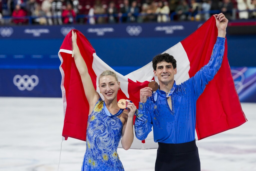 Piper Gilles and Paul Poirer win a bronze medal at the 2026 Milano Cortina Winter Olympics. They are standing on the ice rink holding up the Canada flag behind them while showing off their bronze medals.